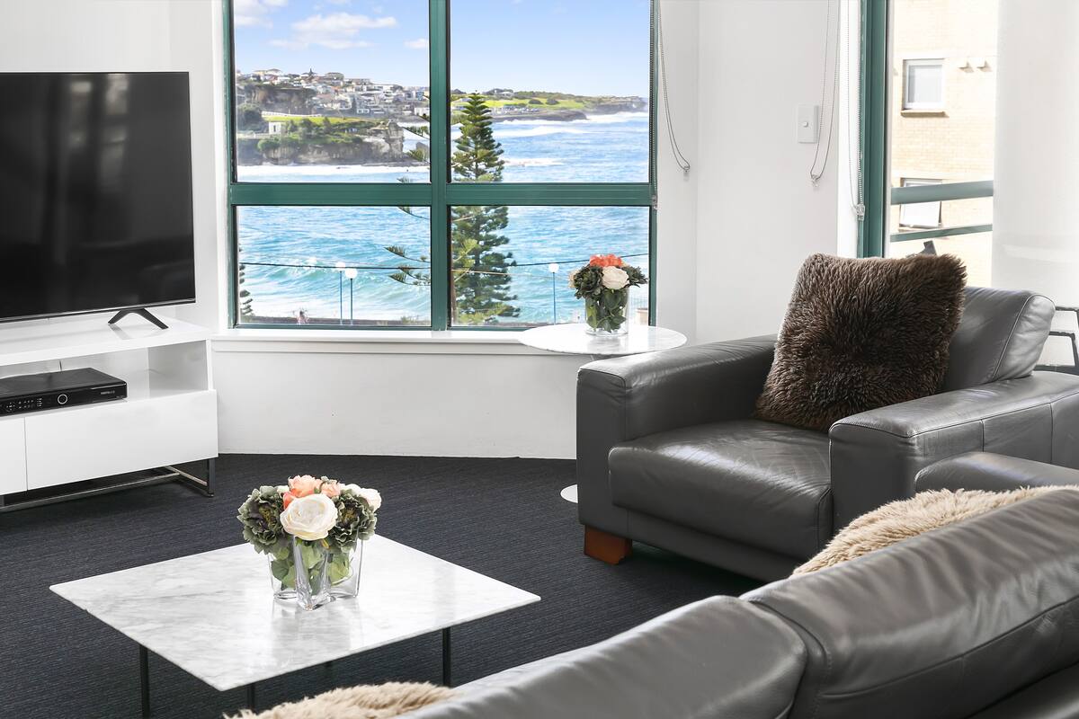 A modern living room with a large window offering a coastal view, a flat-screen TV, a leather sofa, and a coffee table with flowers.