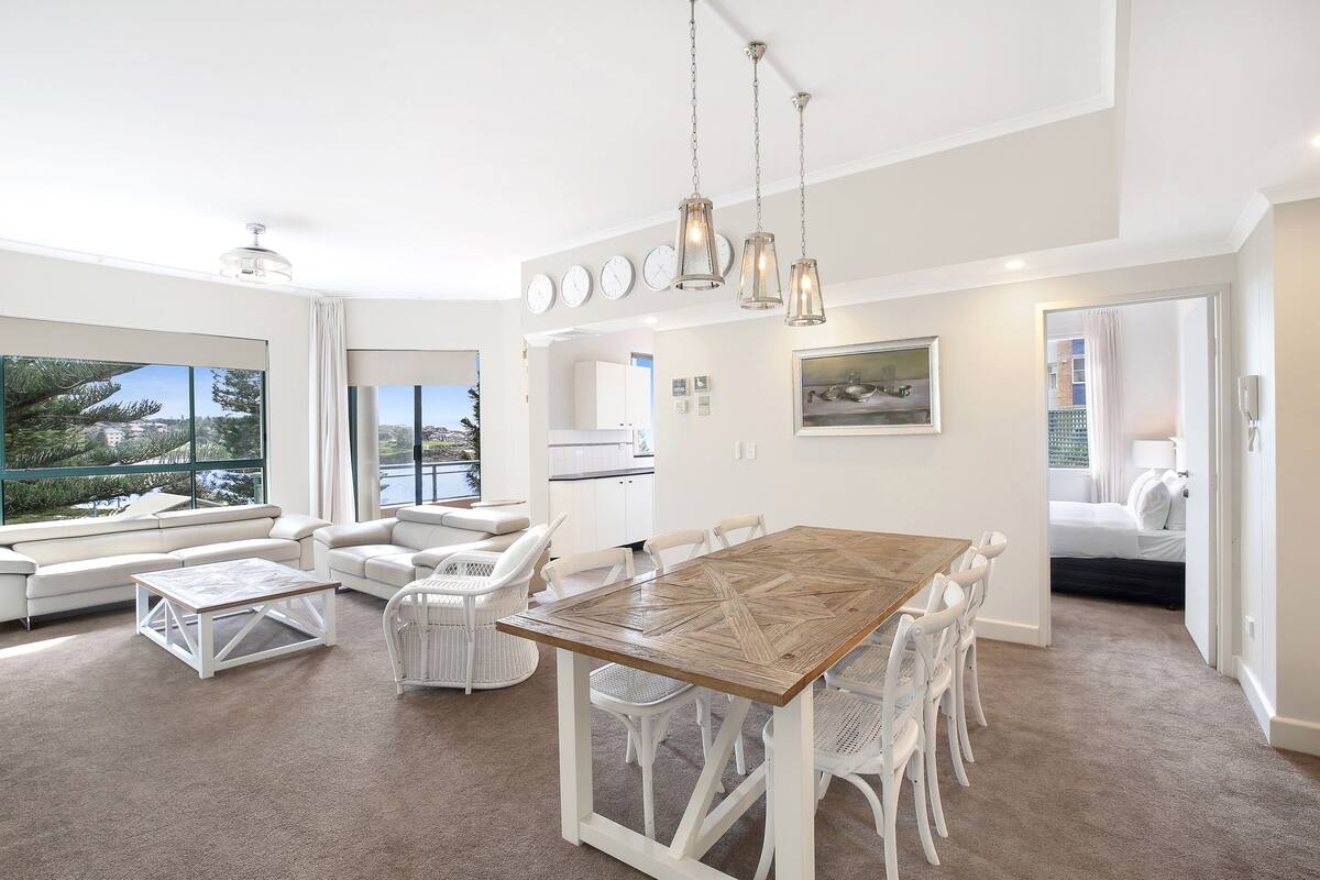 A spacious living area with a wooden dining table, white chairs, and a sofa set. There are large windows offering a view of the outdoors.