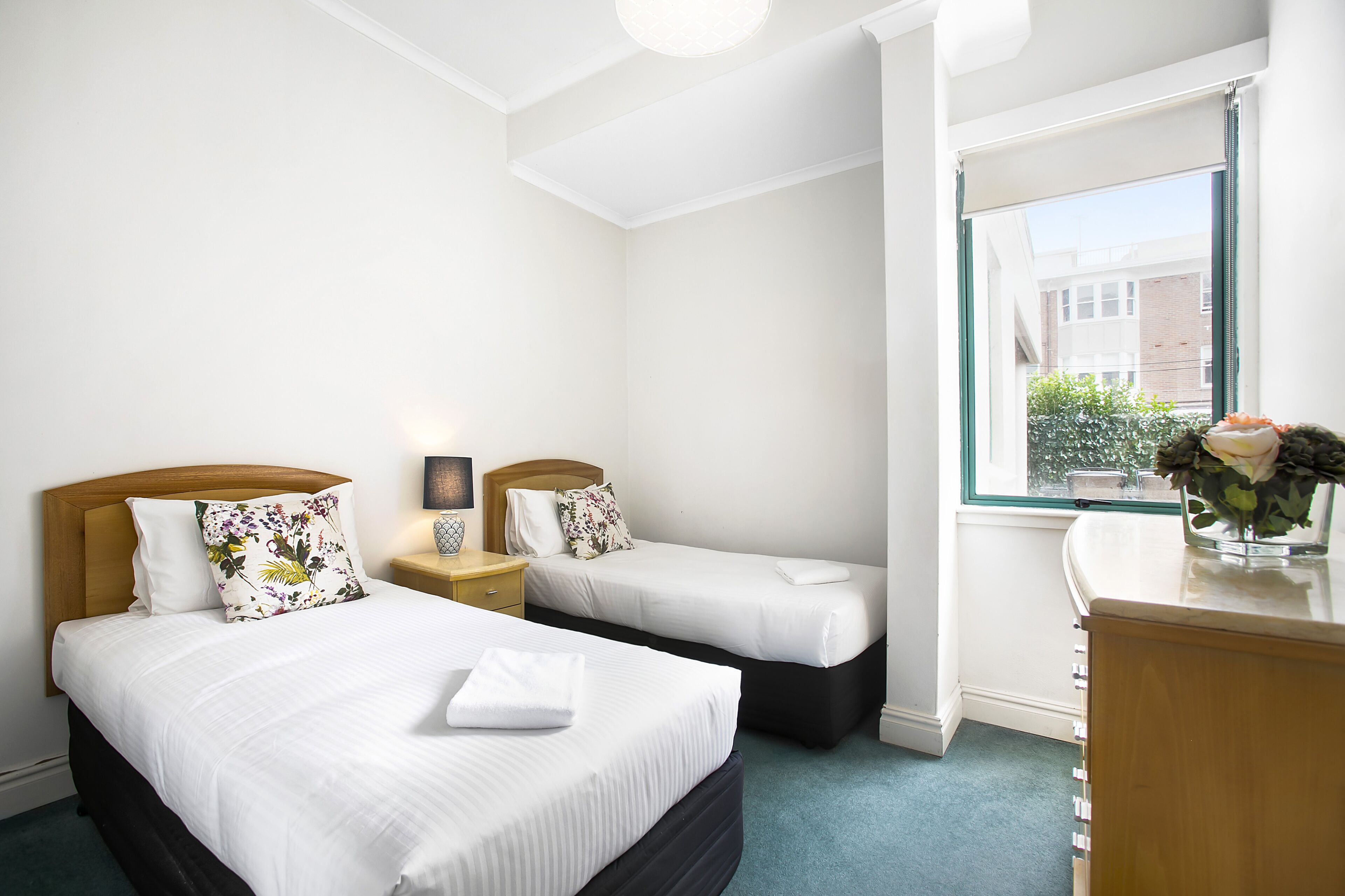 Une chambre d’hôtel avec deux lits, un bureau, une fenêtre donnant sur un beau paysage et une petite table sur laquelle est posé un vase.