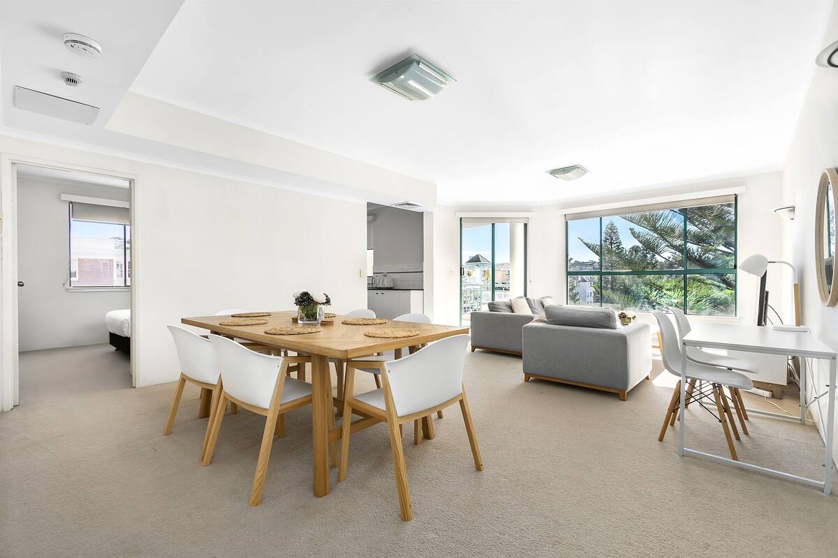 A modern living room with a dining area, large windows, and a view of greenery.