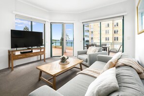 A modern living room with a flat-screen TV, a wooden coffee table, a grey sofa, and a balcony with a view of the sea. - AEA The Coogee View Serviced Apartments (Coogee)