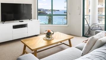 A modern living room with a large flat-screen TV, a wooden coffee table with a vase, and a view of the sea through a large window.
