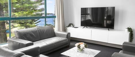 Ett modernt vardagsrum med en grå lädersoffa, ett vitt soffbord med en vas med blommor och en platt-TV placerad på ett vitt skåp.