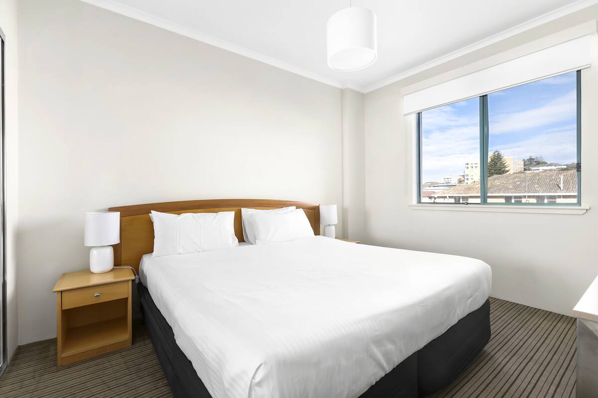 A hotel room with a large bed, a wooden nightstand with a lamp, a window with a city view, and a ceiling light.