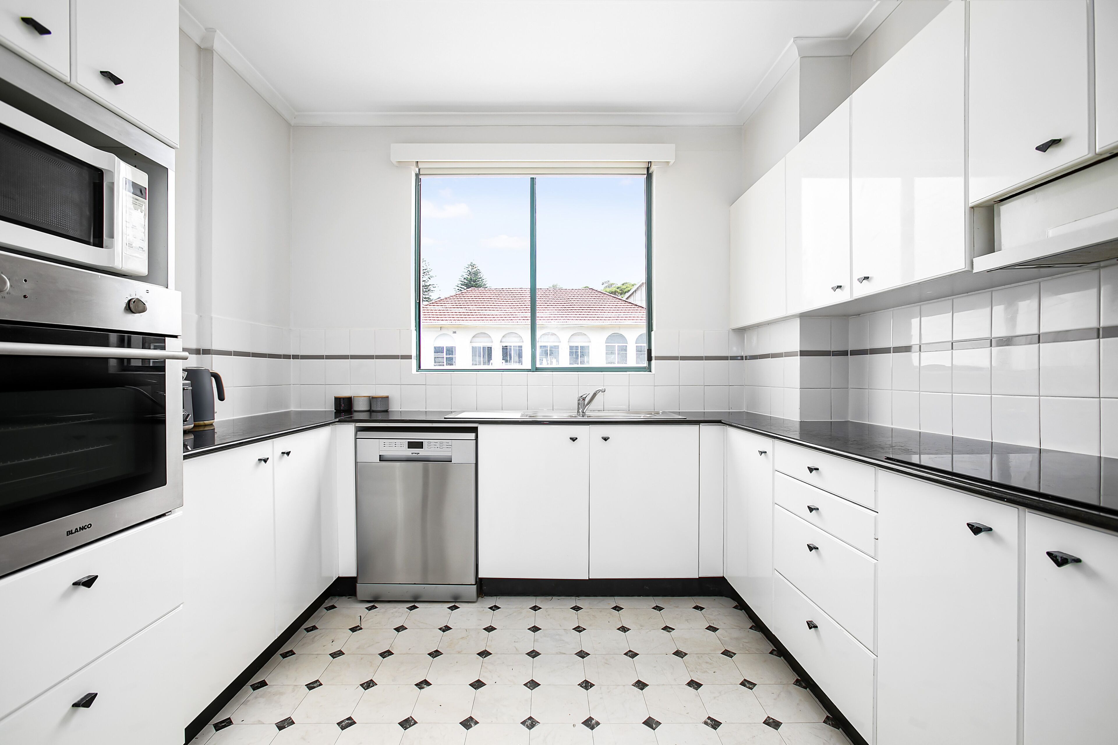 Een moderne keuken met witte kasten, een roestvrijstalen vaatwasser en een raam met uitzicht op een gebouw.