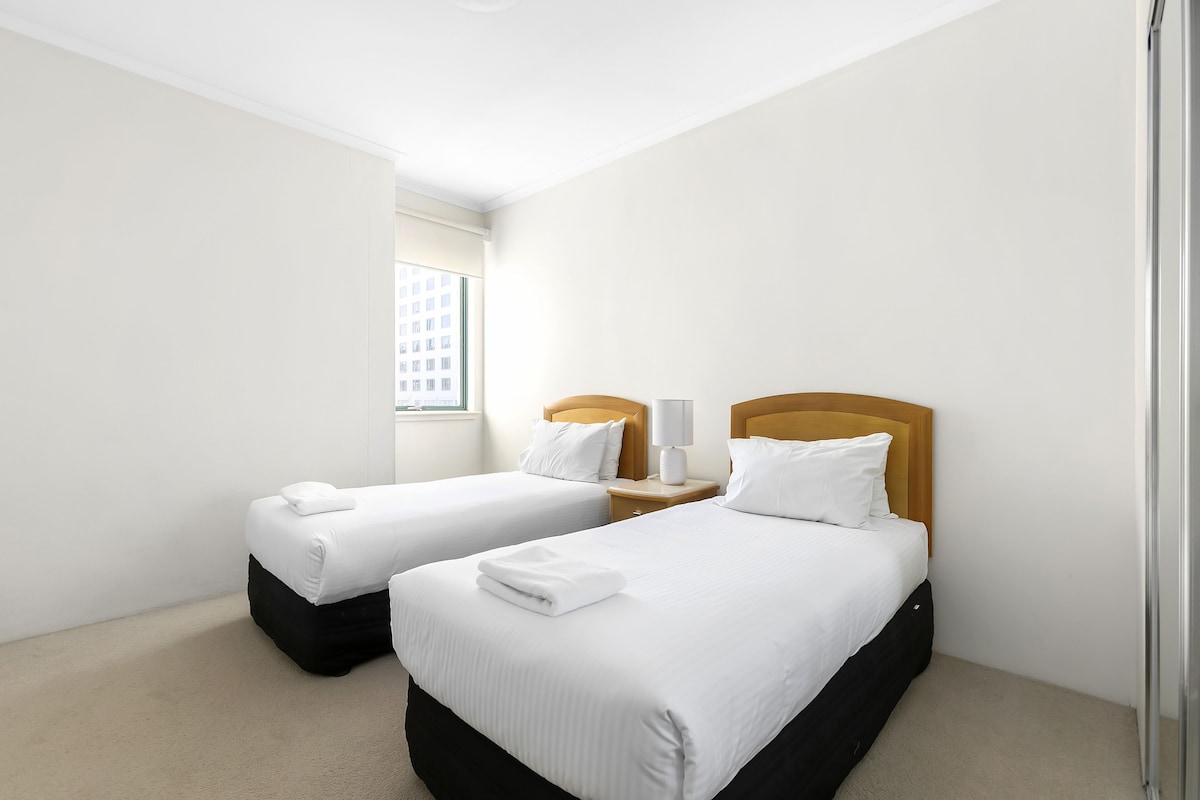 A hotel room with two beds, each with white bedding and a wooden headboard. There is a window providing a view of a building outside.