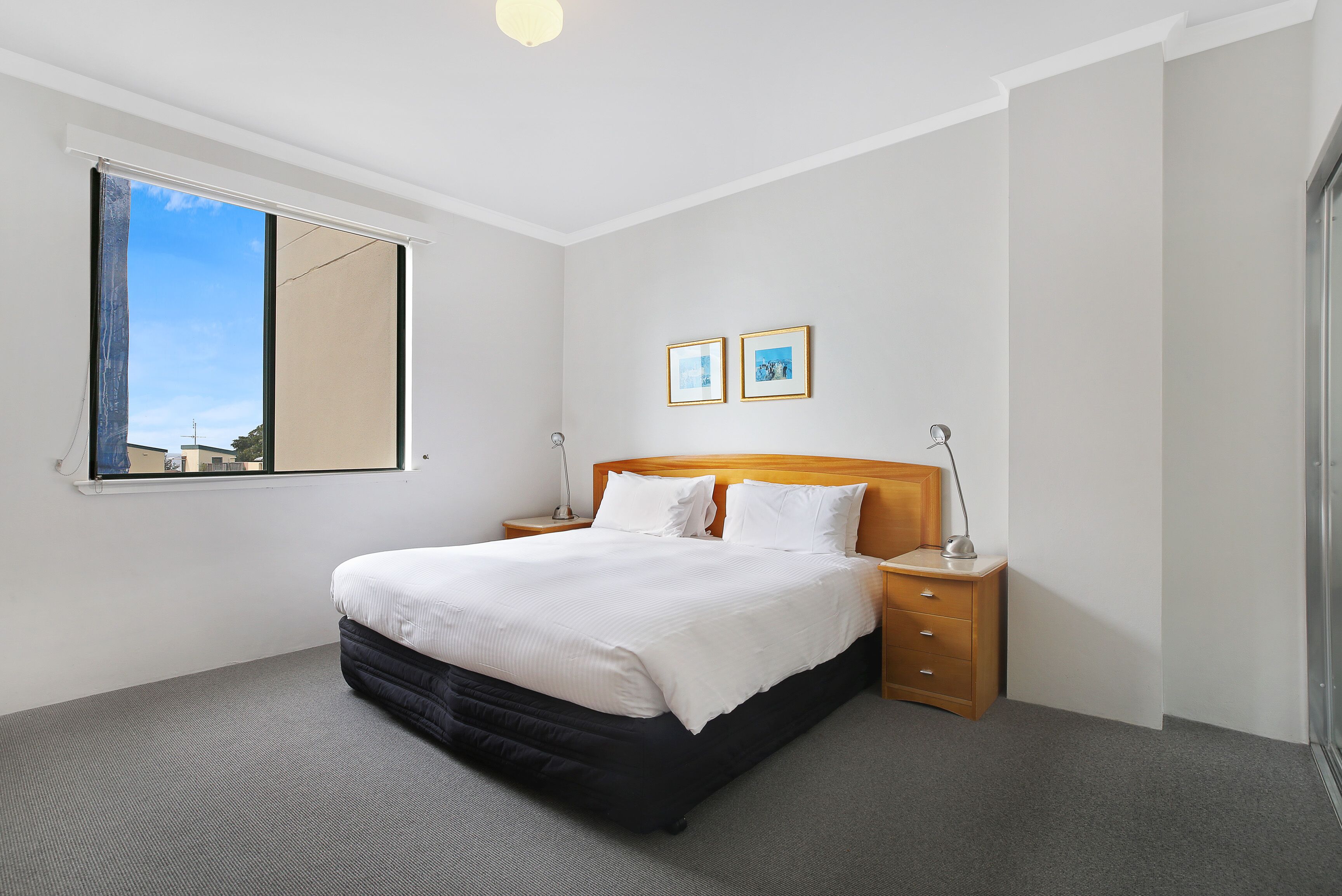 A neatly made bed with a wooden headboard, a bedside table with a lamp, and two framed pictures on the wall.