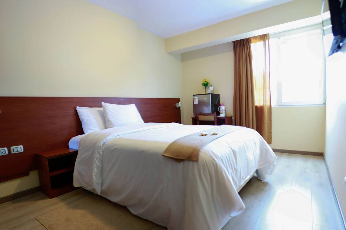 Quarto de hotel com uma cama grande, mesa de cabeceira, escrivaninha com abajur, geladeira e janela com cortinas.
