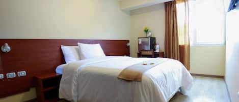 A hotel room with a large bed, a nightstand, a desk with a lamp, a refrigerator, and a window with curtains.