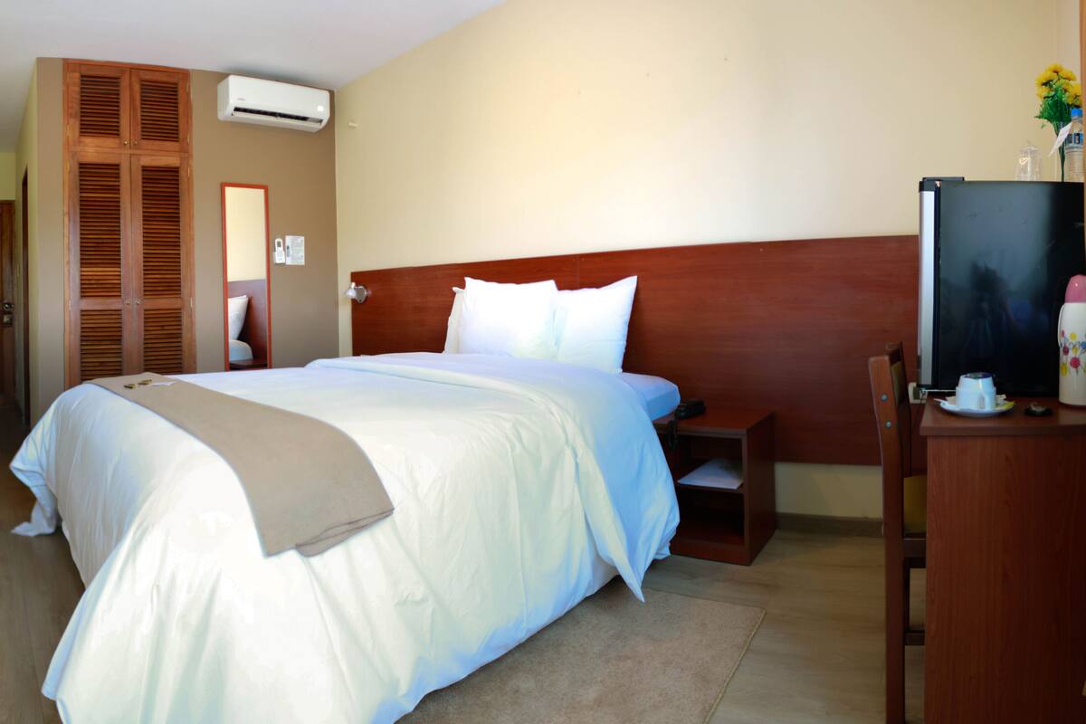 Quarto de hotel com cama grande, cabeceira de madeira, ar condicionado, televisão e uma pequena mesa com xícara e pires.
