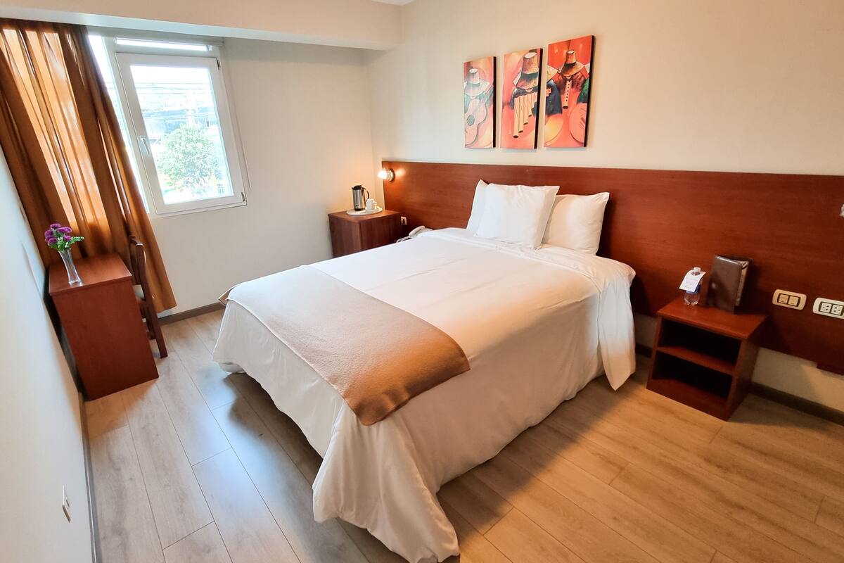 Cama bem arrumada com edredom branco e almofadas, mesa de cabeceira com abajur, criado-mudo com telefone e um quadro na parede.