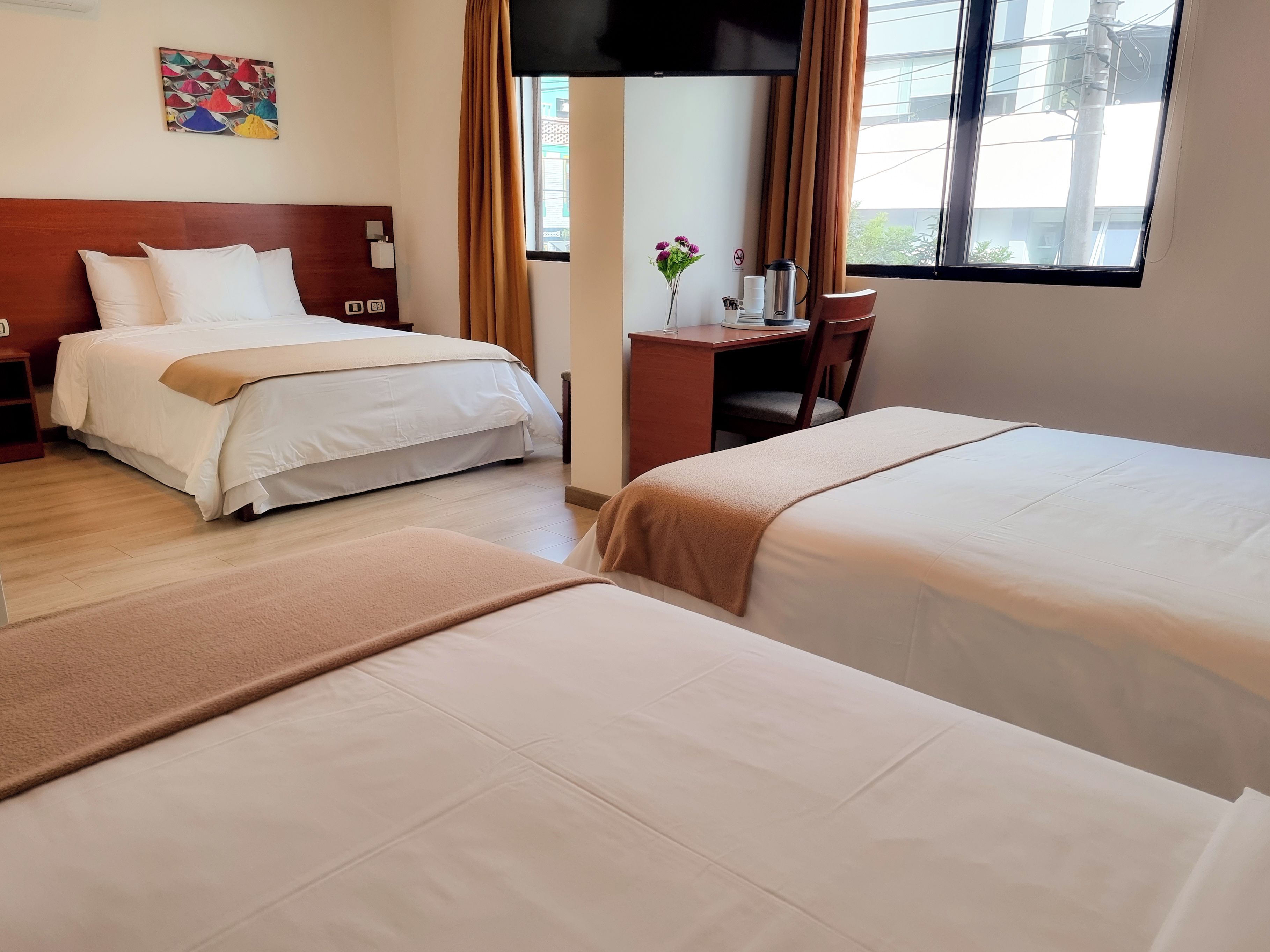A hotel room with two beds, a desk, a chair, a window with curtains, and a wall-mounted TV.