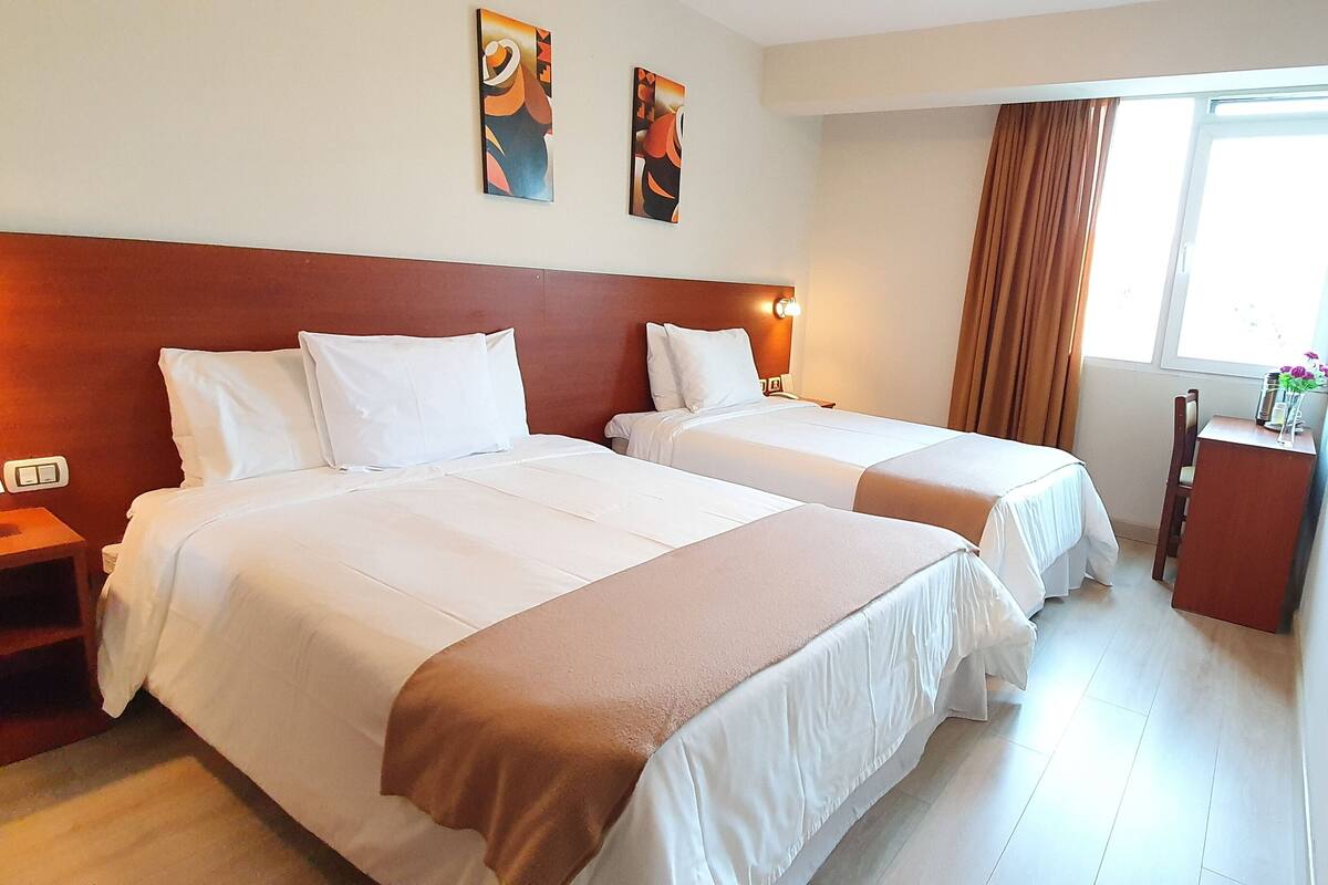 Quarto de hotel com duas camas, uma cabeceira de madeira, mesa de cabeceira com luminária e janela com cortinas.