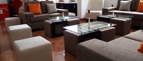 A modern living room with sofas, ottomans, and a glass coffee table. There are two computer monitors on a desk.