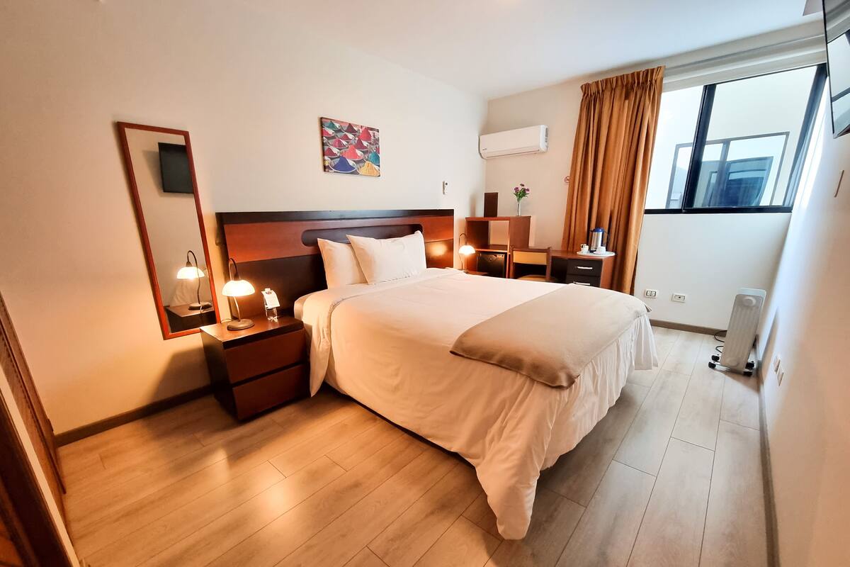 Quarto de hotel com uma cama grande, mesas de cabeceira com luminárias, uma escrivaninha com cadeira, uma pequena mesa com um vaso e uma janela com cortinas.