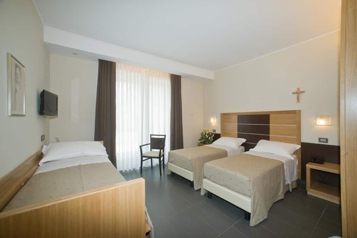 Habitación de hotel con dos camas individuales, una silla, una mesita, un televisor y una cruz en la pared.