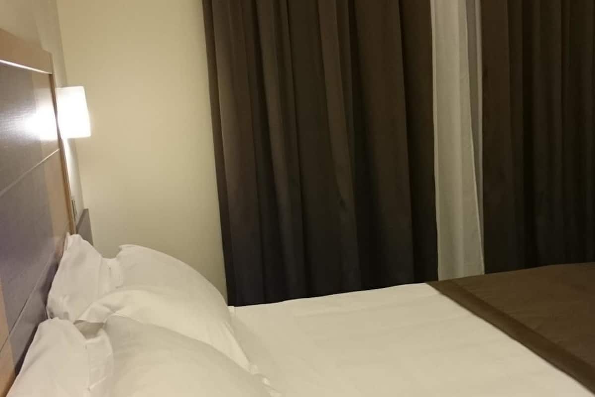 Una cama bien hecha con sábanas y almohadas blancas, una lámpara de noche y una ventana con cortinas oscuras.