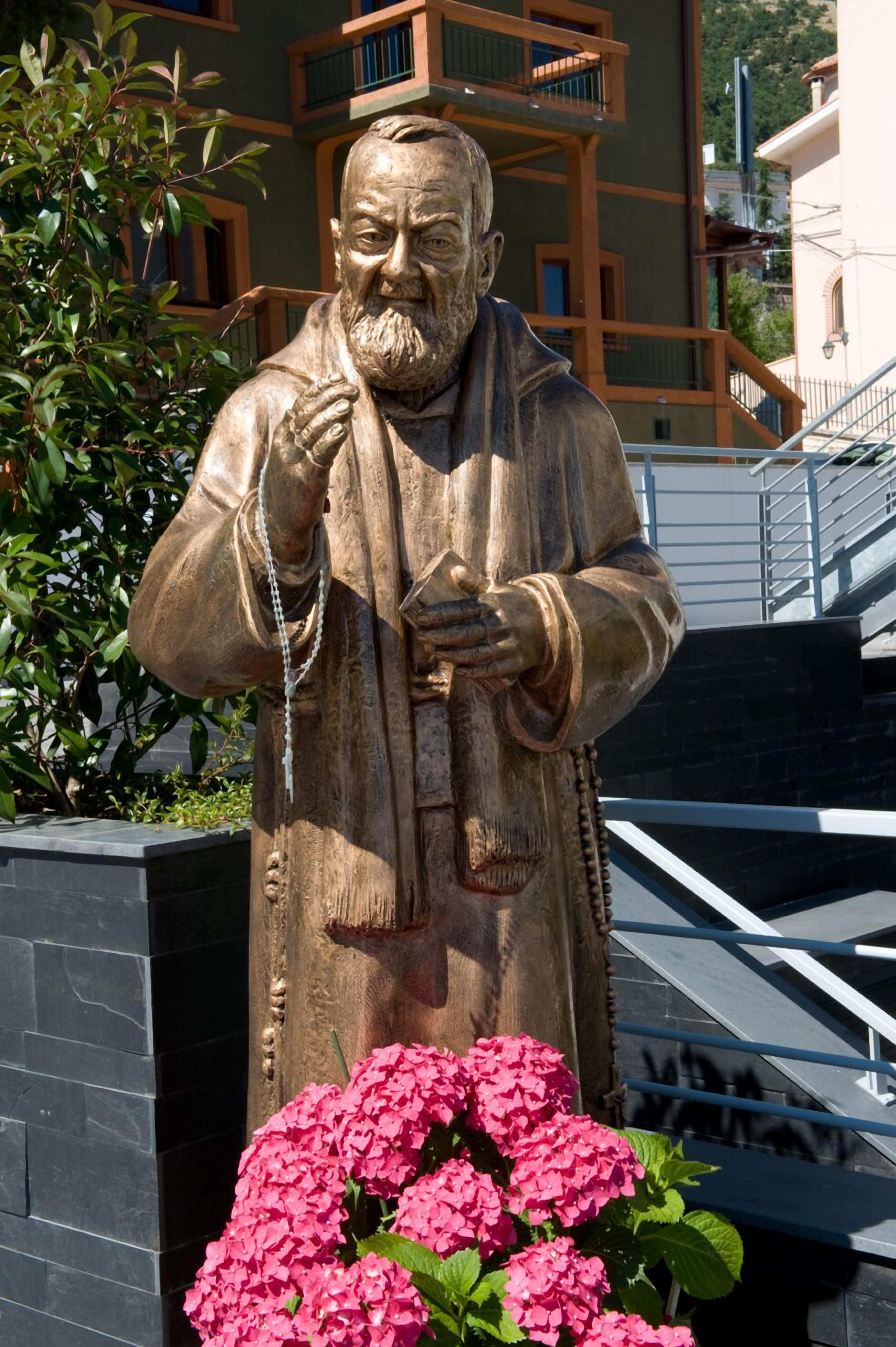 Estátua de uma pessoa vestida com traje tradicional, segurando um livro, com uma escadaria e edifícios ao fundo.