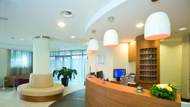 A modern reception area with a curved wooden desk, a seating area with a round couch, and a large potted plant. - Vittoria (San Giovanni Rotondo)