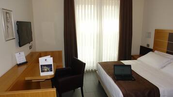 Une chambre d’hôtel avec un lit, un bureau avec un ordinateur portable, une chaise, une télévision et une fenêtre avec des rideaux.