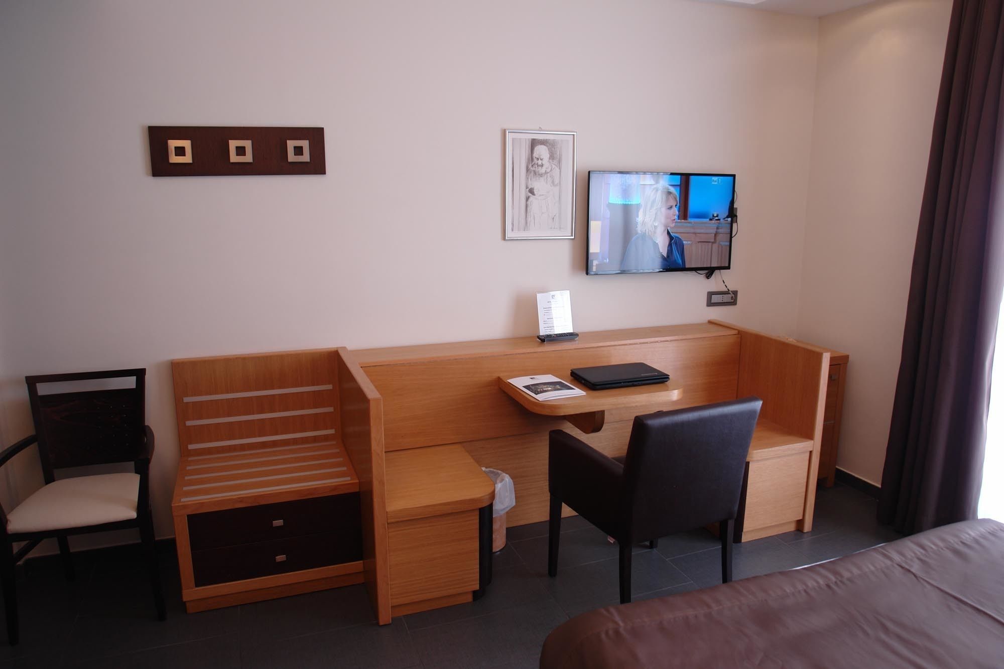 A hotel room with a wooden desk, a chair, a bed, a TV, and a framed picture on the wall.