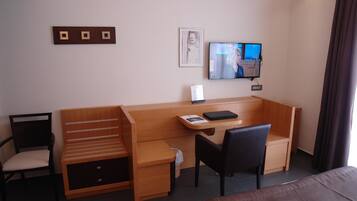 A hotel room with a wooden desk, a chair, a bed, a TV, and a framed picture on the wall.