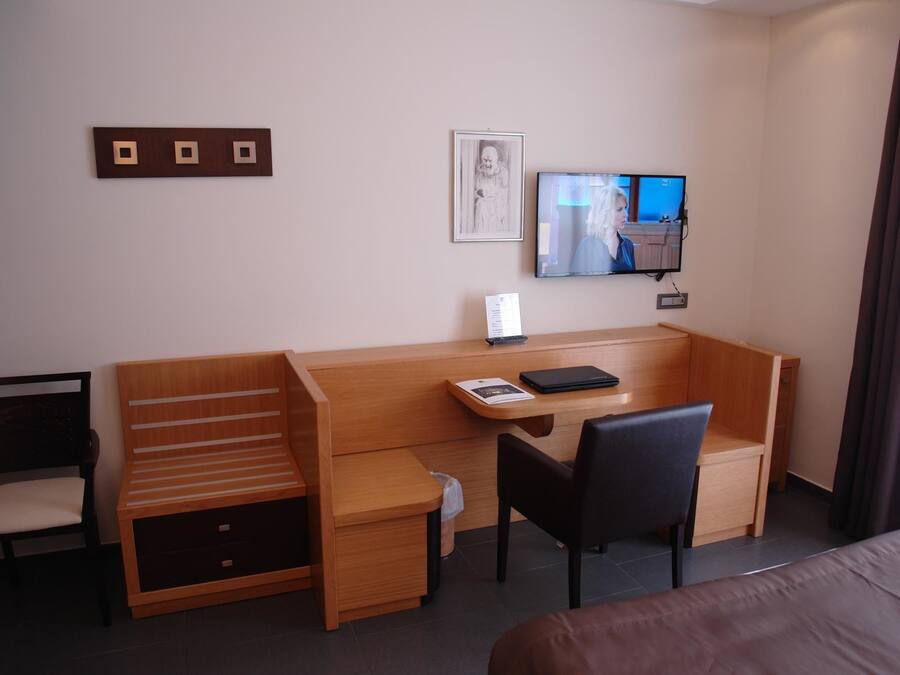 A hotel room with a wooden desk, a chair, a bed, a TV, and a framed picture on the wall.