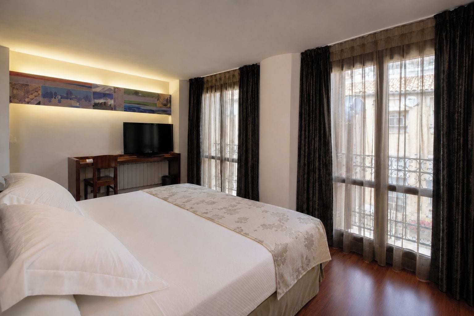 Habitación de hotel con una cama, un televisor sobre un mueble bajo y grandes ventanales con cortinas.