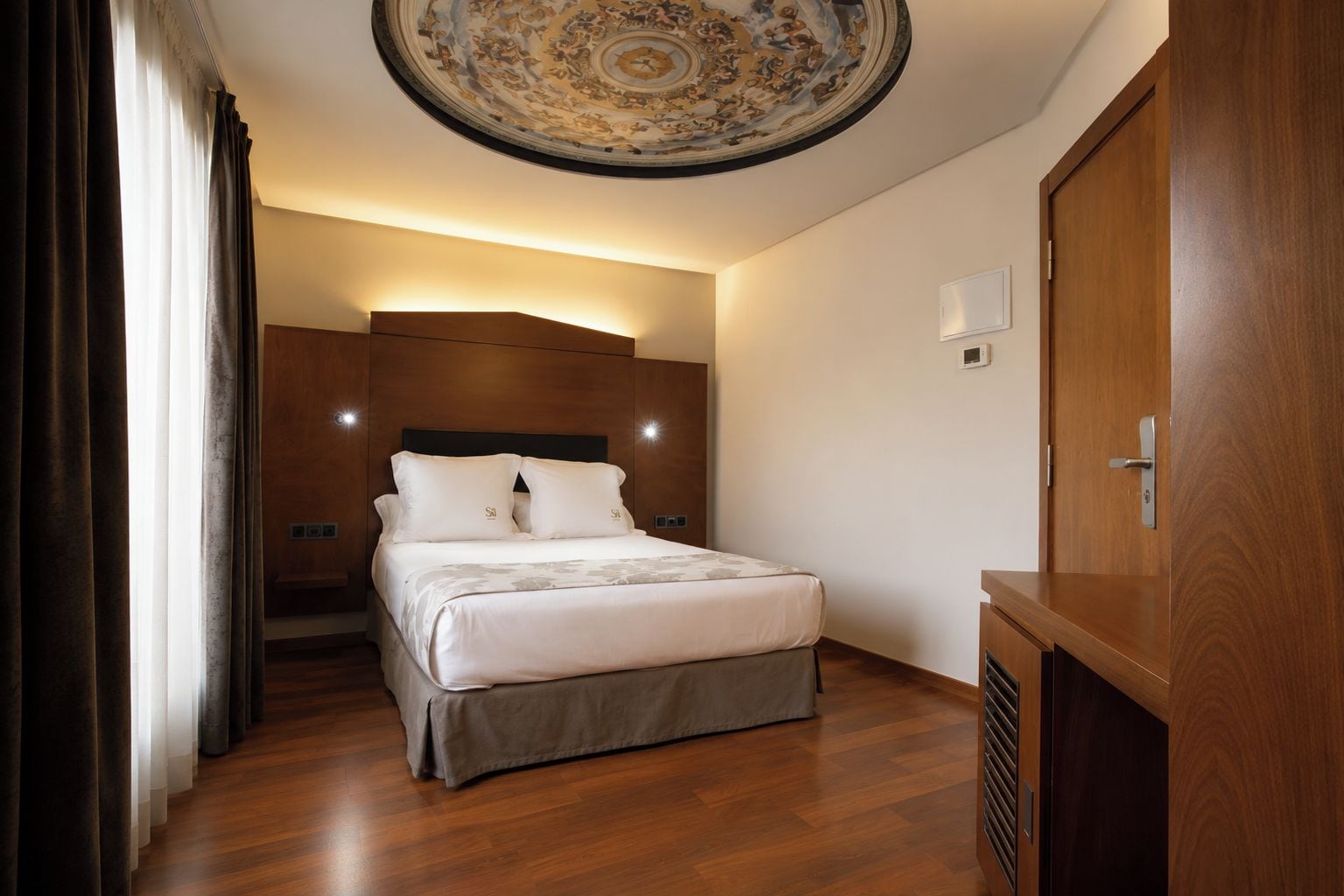 A hotel room with a bed, wooden headboard, a ceiling with a decorative design, a wooden wardrobe, and a window with curtains.