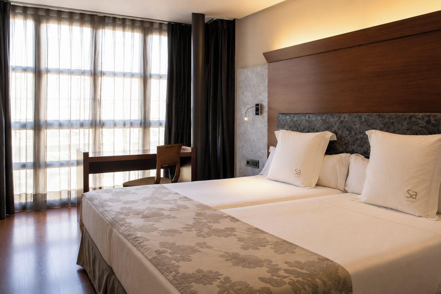 Een hotelkamer met een bed, een bureau en een stoel. Er is een raam met gordijnen, een wandlamp en een hoofdeinde.