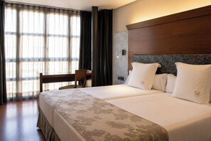 A hotel room with a bed, a desk, and a chair. There is a window with curtains, a wall-mounted light, and a headboard.