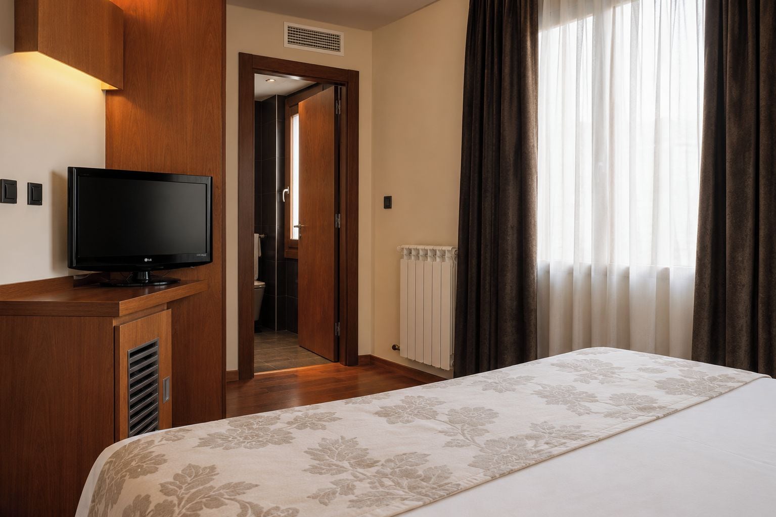 Habitación de hotel con cama, un televisor sobre un mueble de madera, un baño visible a través de una puerta abierta y una ventana con cortinas.