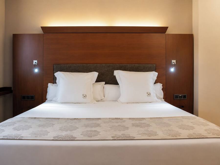 A hotel room with a wooden headboard, two white pillows with a logo, and a floral-patterned bedspread.