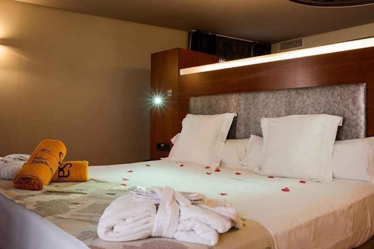 Una habitación de hotel con cama, ropa de cama blanca, cabecera y toalla sobre la cama.