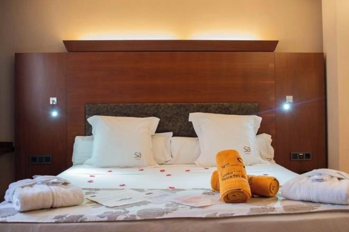 Una cama bien hecha con sábanas blancas y toallas enrolladas, un cabecero de madera y una mesita de noche con una lámpara.