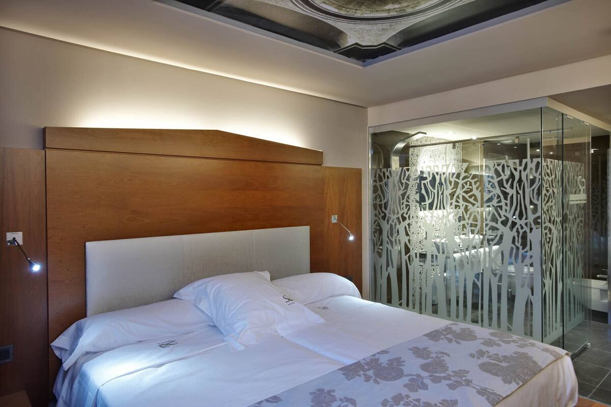 Un dormitorio moderno con cabecera de madera, cama con ropa de cama blanca y separador de vidrio con diseños blancos intrincados.
