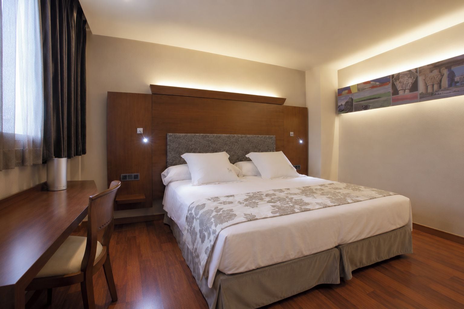 A hotel room with a bed, bedside table, chair, and a painting on the wall.