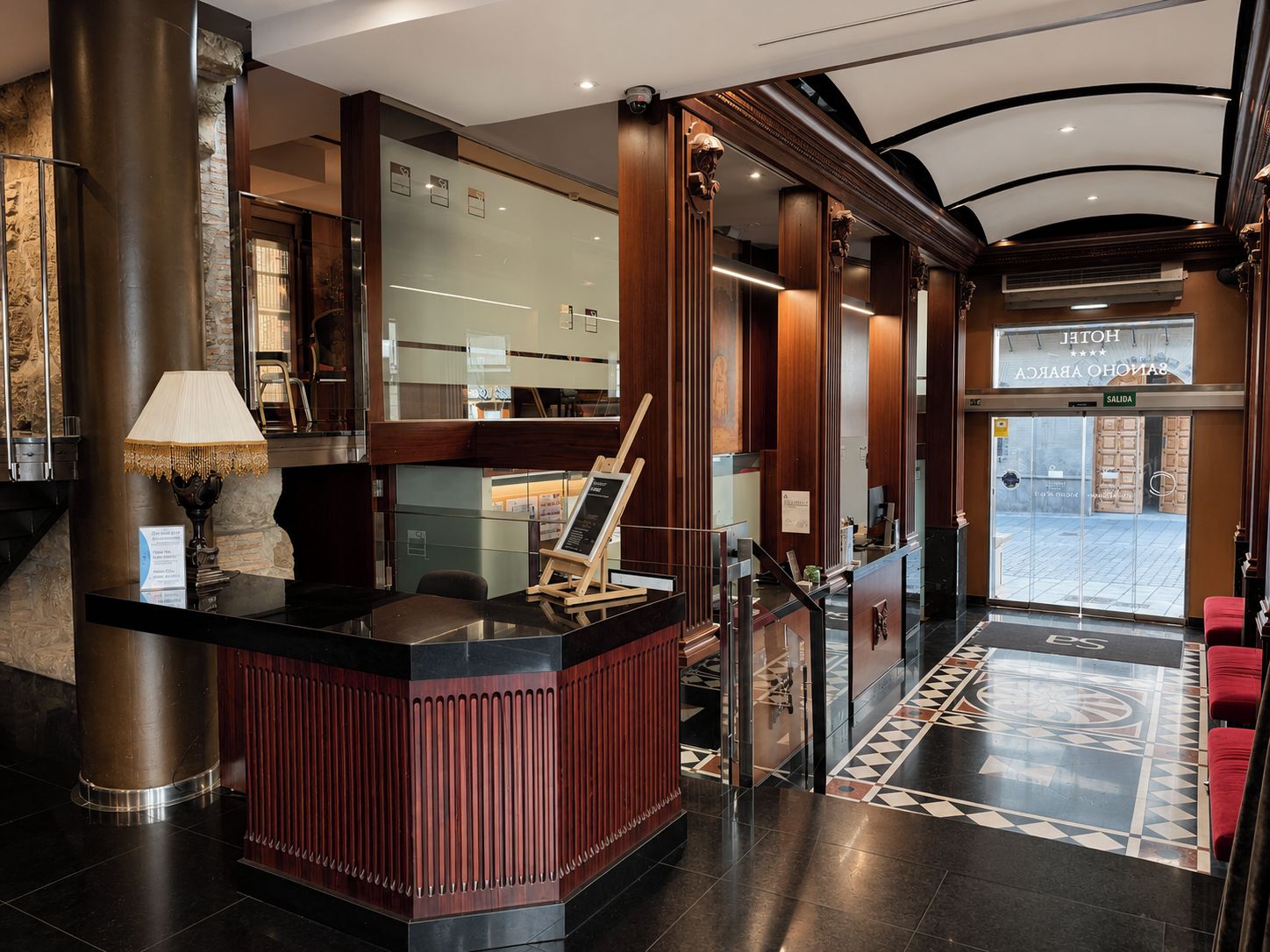 A hotel lobby with a reception desk, a red accent wall, a large mirror, and a staircase leading to the upper floor.