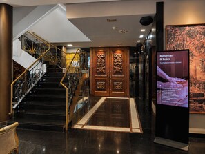 A hotel lobby with a staircase, a reception desk, and a digital information board.