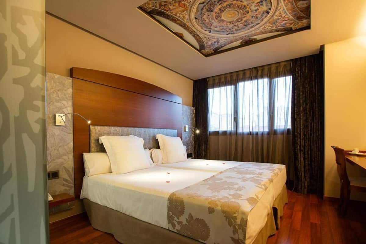 Una cama doble con cabecera, una mesita de noche, un escritorio y un ventanal con cortinas.
