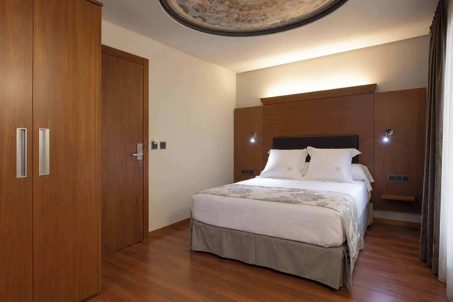 Habitación de hotel con un armario de madera, una cama con ropa blanca y una lámpara fijada a la pared.