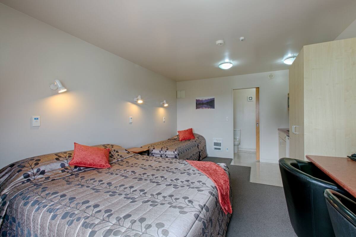 Une chambre d’hôtel avec deux lits, un bureau et une chaise. Des lampes sont fixées au mur, et une porte donne accès à une autre pièce.
