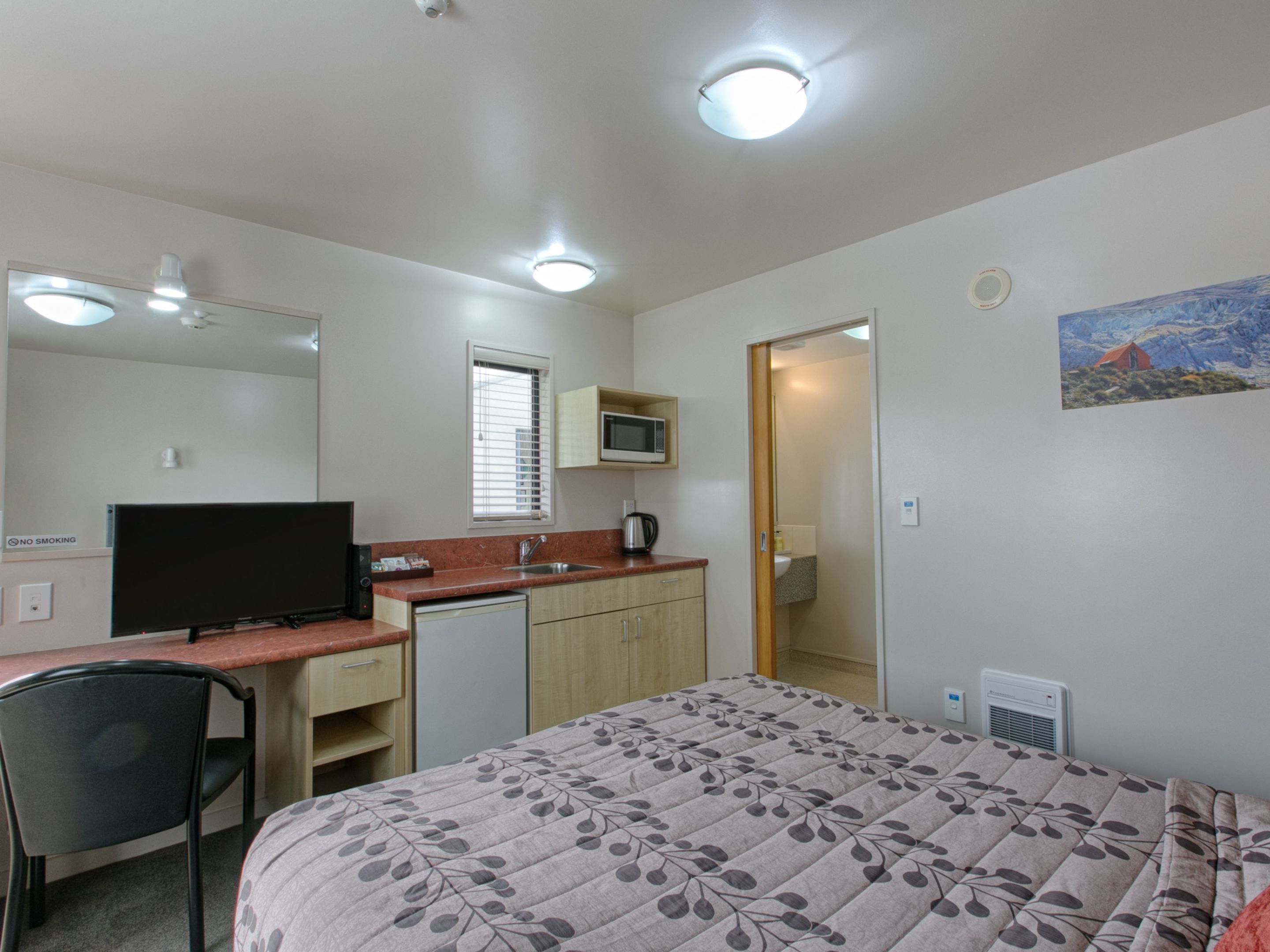 A compact room with a bed, a desk, a chair, a microwave, and a small kitchenette.