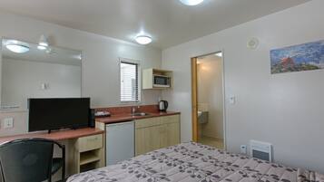 A compact room with a bed, a desk, a chair, a microwave, and a small kitchenette.