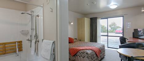 Quarto de hotel com chuveiro, cama com manta vermelha, escrivaninha com cadeira e vista para o exterior por uma porta deslizante.