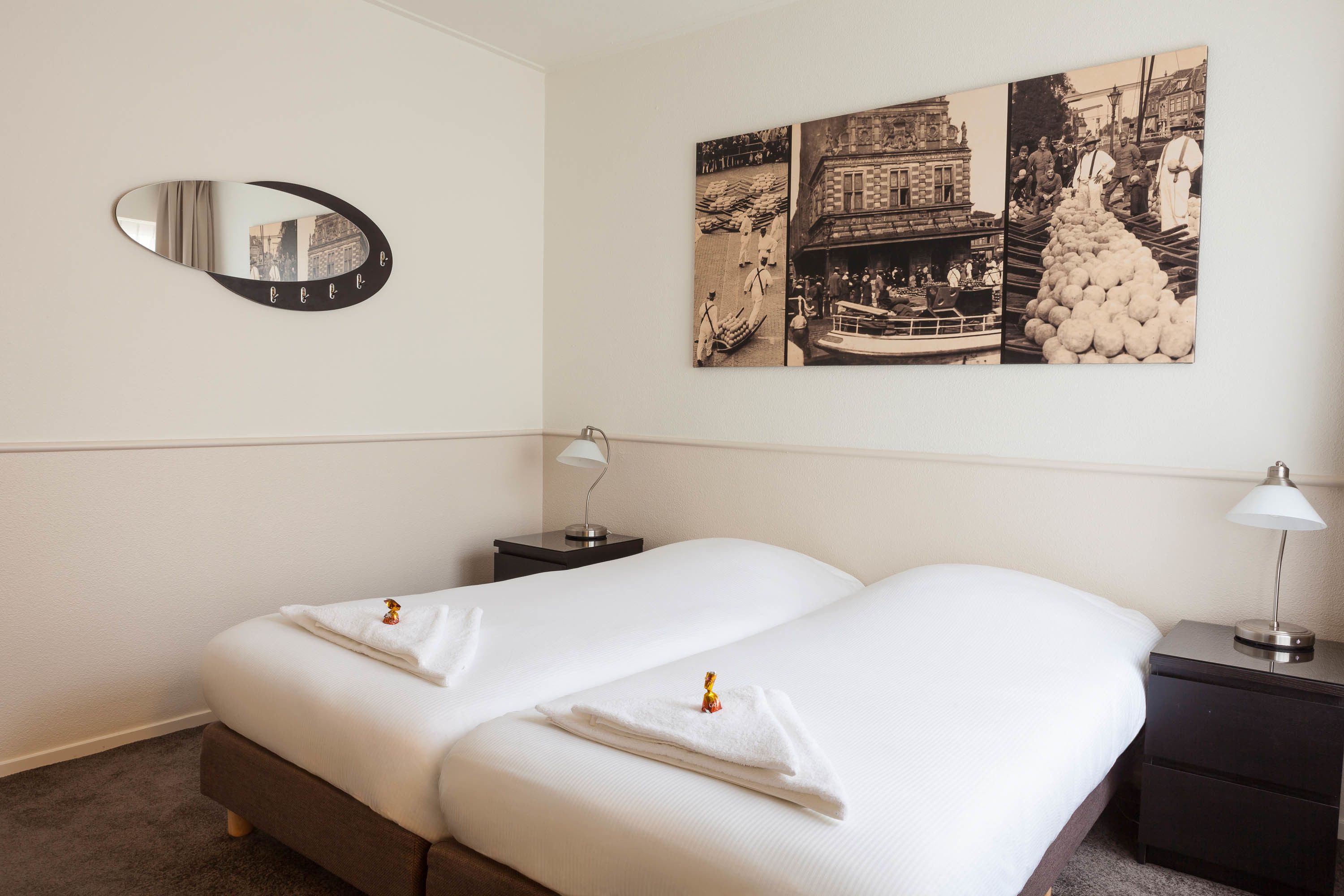 A hotel room with a double bed, a nightstand, a lamp, a mirror, and a wall with a black and white photograph.