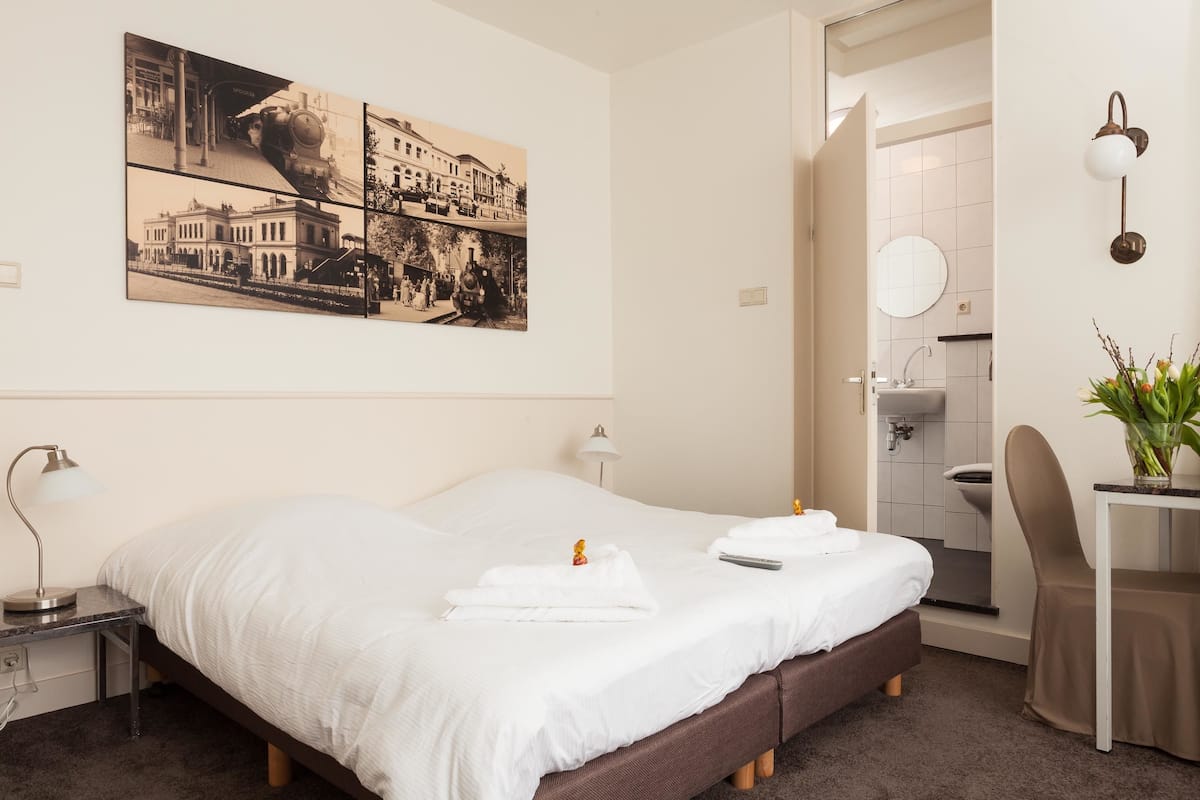 A hotel room with a large bed, a desk with a lamp, a chair, a potted plant, and a wall-mounted mirror.
