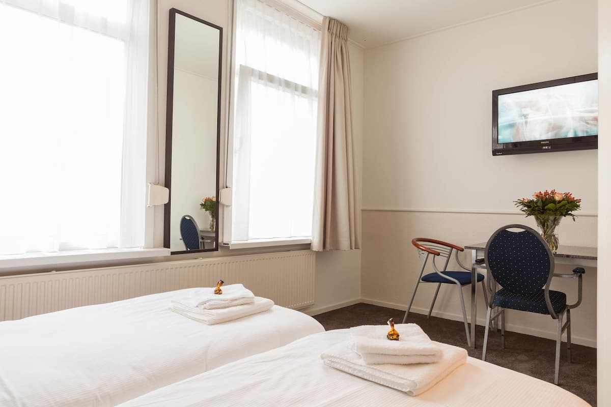 A hotel room with two beds, a desk with a chair, a TV on the wall, and a window with curtains.