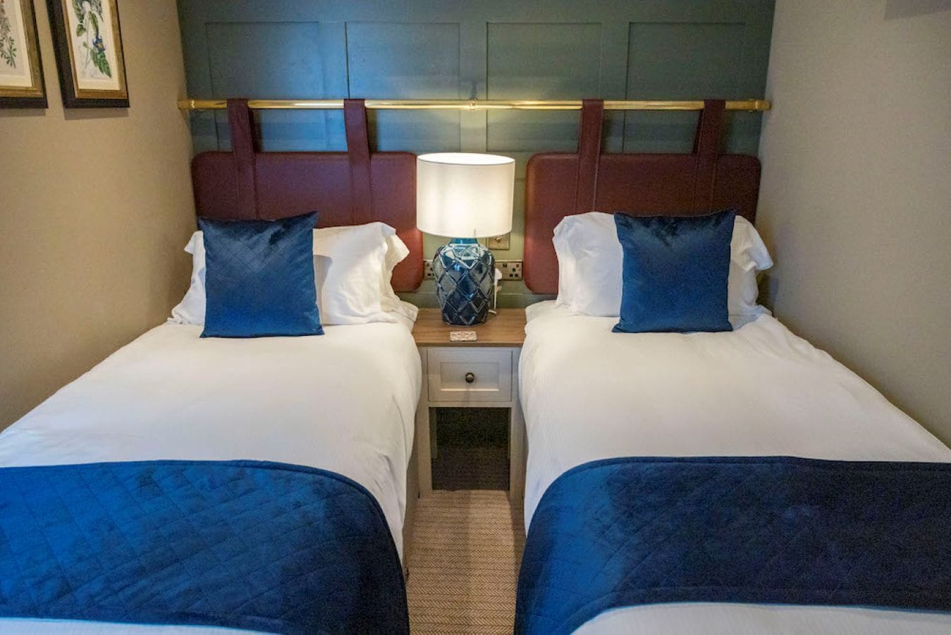 A hotel room with two beds, a nightstand with a lamp, and a headboard with a decorative panel.