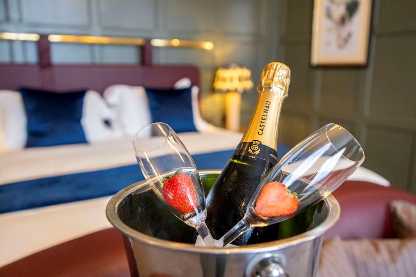 A bottle of Castelnau champagne in a metal bucket with two glasses, one with a strawberry.
